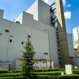 A modern power plant with large industrial buildings, a tall chimney, and a cooling tower, surrounded by green landscaping and trees under a blue sky.