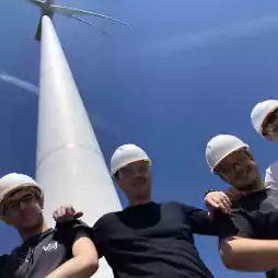 Five people wearing white safety helmets and glasses stand together smiling in front of a tall wind turbine on a clear, sunny day.