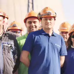 A group of industrial workers wearing orange safety helmets, safety glasses, and branded clothing stand together outside. The workers are smiling and appear to be part of the AES Bulgaria company, as indicated by the logo in the top right corner.