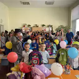 A lively classroom scene with young children standing at their desks, holding colorful balloons and backpacks. Two adults are engaging the class, while parents and teachers observe from the back. The desks are decorated with flowers and school supplies, creating a festive atmosphere.