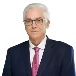 Older man with white hair and glasses wearing a navy blue suit, white shirt, and red patterned tie, standing in a modern office environment with blurred background.