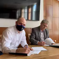 Two men sitting at a wooden table signing documents in an office setting. One man wears a white shirt and a black face mask, while the other wears a gray blazer. A large TV is mounted on the wall behind them and sunlight enters through a window.