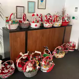 Several decorated baskets with red and white ornaments are displayed on and in front of a wooden cabinet in an office setting. The baskets contain handmade crafts and festive items, with certificates and a trophy on the wall above.