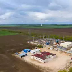 Aerial view of a renewable energy facility with wind turbines in the background, a substation with electrical equipment, and several buildings surrounded by farmland under a cloudy sky.