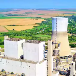 Aerial view of a large power plant with a cooling tower and several industrial buildings surrounded by green fields and farmland under a clear blue sky.