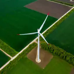 Aerial view of a large wind turbine with three blades standing in the middle of green agricultural fields, bordered by dirt roads and trees.