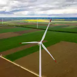 Aerial view of large wind turbines on green and brown agricultural fields under a cloudy sky, illustrating renewable energy production in a rural landscape.
