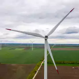A large wind turbine stands in the foreground of a green agricultural landscape with several other wind turbines spread across the fields under a cloudy sky.