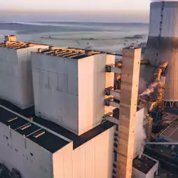 Aerial view of a large industrial power plant with white buildings, cooling tower, and steam rising from several points, set against a rural landscape at sunrise.