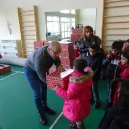 An adult hands a wrapped gift box to a young girl in a pink coat while several children and adults watch in a gymnasium with stacked gift boxes in the background.