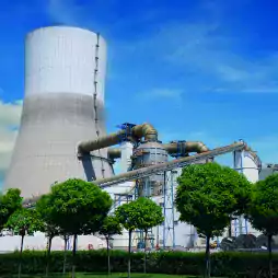 A large industrial cooling tower with pipes and structures is seen behind a row of green trees under a blue sky with scattered clouds.