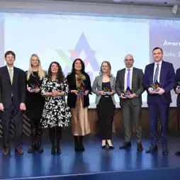 Nine people in formal attire stand on a stage holding colorful awards during a ceremony. A large screen behind them displays text and a geometric logo, indicating an awarding event in Sofia on December 15, 2021.