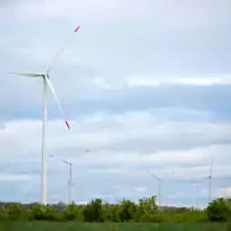 Several wind turbines stand in a green field with trees under a cloudy sky, generating renewable energy.