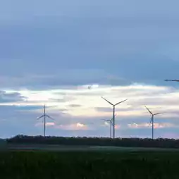 Several wind turbines stand in a green field under a cloudy sky at sunset, generating renewable energy.