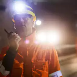 A worker wearing an orange safety jacket and a yellow helmet with a headlamp communicates via walkie-talkie inside an underground tunnel. Bright lights and two other workers in similar safety gear are visible in the background.