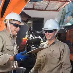Two male workers wearing safety helmets and sunglasses stand next to an orange truck with its hood open. One man holds a clipboard and both are smiling, suggesting a discussion or inspection at a work site.