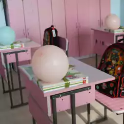 Pink classroom with several desks, each with a balloon, a stack of books tied with ribbon, and a colorful children's backpack. The classroom has pink cabinets and light wood flooring, creating a cheerful and welcoming environment.