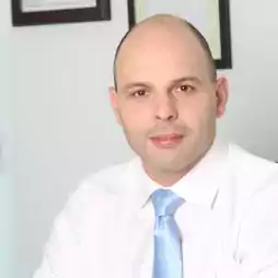 A bald man in a white shirt and light blue tie sits in an office chair, looking at the camera with a neutral expression. Framed certificates are visible on the white wall behind him.