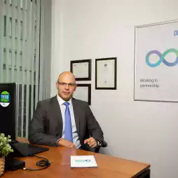 A man in a business suit sits at a desk in a modern office with a computer, potted plant, certificates on the wall, and a poster that reads 'Working in partnership' with the aes logo.