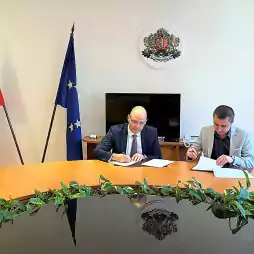 Two men sit at a conference table signing documents in an office. The Bulgarian and European Union flags are displayed in the background, along with the Bulgarian coat of arms on the wall. Green plants decorate the edge of the table.