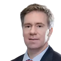 Portrait of a man with light brown hair wearing a dark suit, light blue shirt, and patterned tie, against a white background.