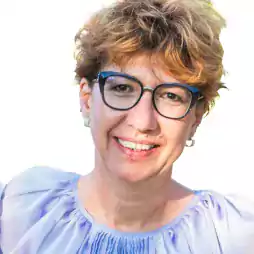 Smiling woman with short curly light brown hair and glasses wearing a light blue blouse, photographed against a white background.