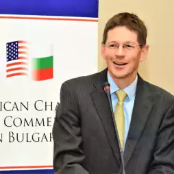 A man in a suit and tie speaks at a podium with a microphone, standing in front of a banner displaying the American and Bulgarian flags and the text 'American Chamber of Commerce in Bulgaria.'