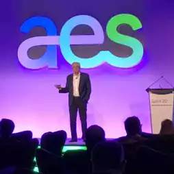 A man in a suit is giving a presentation on stage in front of a large colorful 'aes' logo, with an audience seated in the foreground and a podium labeled 'Spark 20' to the right.