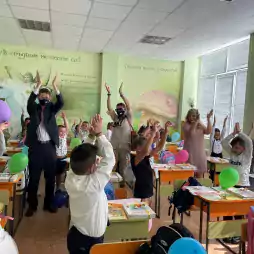 Children and teachers in a brightly lit classroom with colorful balloons raise their hands together in an engaging activity. The classroom walls are decorated with murals, including a unicorn and inspirational text, creating a cheerful learning environment.