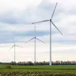 Three large wind turbines stand in a green field under a cloudy sky, with trees lining the horizon. The turbines are used for generating renewable energy from wind power.