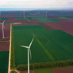 Aerial view of a wind farm with several wind turbines spread across green and brown agricultural fields under a cloudy sky.