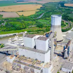 Aerial view of a large power plant with a prominent cooling tower surrounded by green fields, roads, and industrial buildings, illustrating energy infrastructure in a rural landscape.