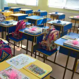 A brightly lit classroom with yellow and blue desks, each desk has a colorful backpack, books, and a pink decorative item neatly arranged, ready for students to arrive.