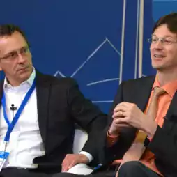 Two men in business attire are seated and engaged in a panel discussion. One man is listening attentively while the other is speaking and gesturing with his hands. Both wear suits and microphones, with a blue background featuring abstract lines.