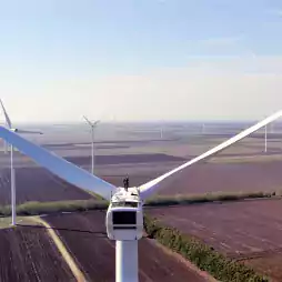 A person stands on top of a large wind turbine in a field, with multiple wind turbines visible in the background under a clear sky, showcasing renewable energy and modern technology.