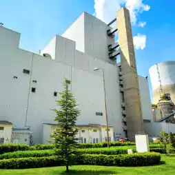 Modern industrial power plant with large white buildings, a tall chimney, and a cooling tower, surrounded by green landscaped grass and trees under a blue sky.