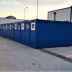 Row of blue portable office containers with small windows and doors, positioned in a parking lot near modern white buildings during daylight.