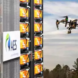 A drone flies near an AES energy storage facility outdoors, with battery racks and company logos visible on the left, and trees under a partly cloudy sky in the background.