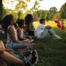 A group of children sit on grass in a park, attentively watching something in the distance on a sunny day. Orange safety cones are visible in the background, and the scene is shaded by trees.